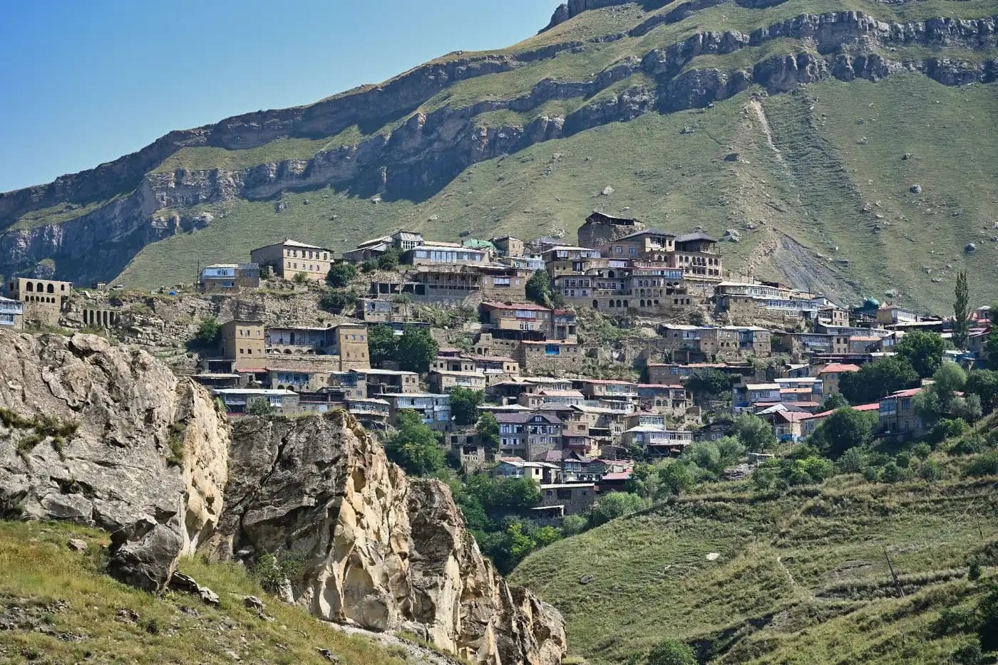 Панорама аула Чох в Дагестане летом — жёлтая каменная застройка на скалистом склоне среди зелёных гор