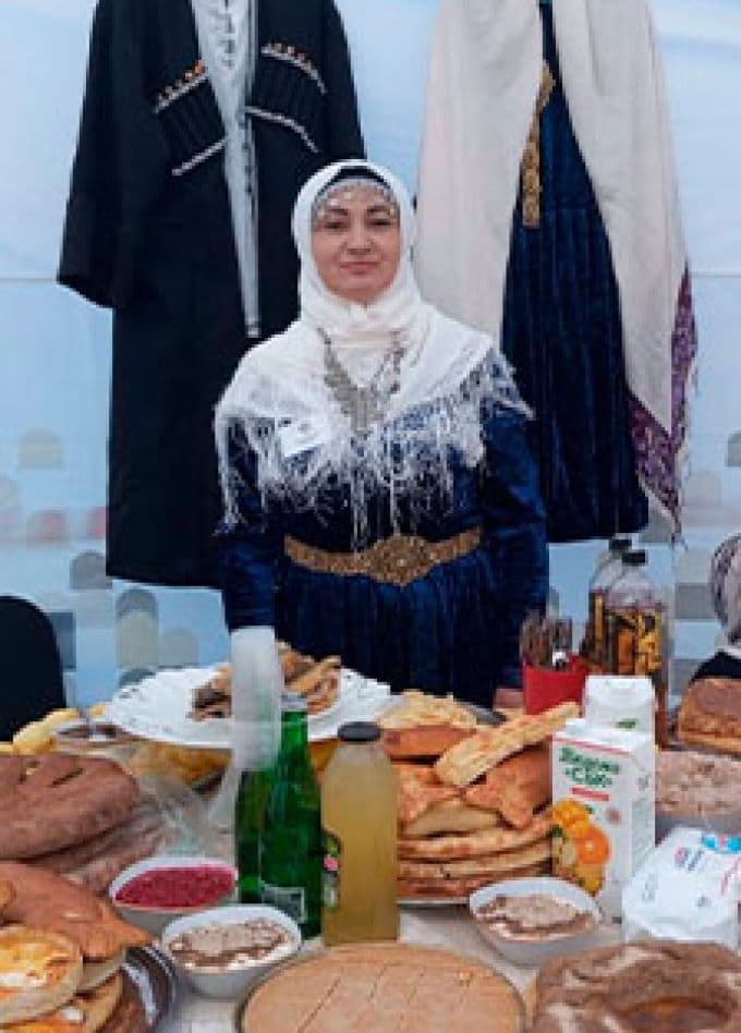 Женщина в национальном костюме у стола с выпечкой и другими блюдами — иллюстрация из афиши программы «Вкус народов России»